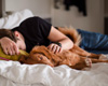 Niedergeschlagene Person im schwarzen Hemd liegt auf dem Bett und Hund liegt bei ihr um zu trösten.